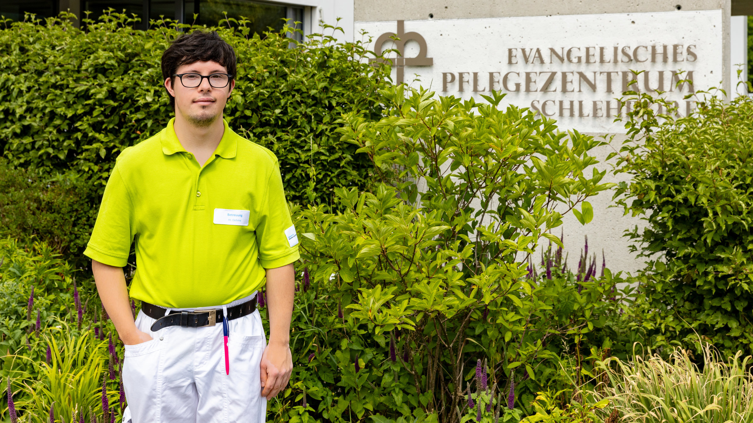 "einfach Mensch - Moritz Oehme - Ich arbeite gerne im Seniorenheim": Moritz Oehme  (22) steht im Freien vor Büschen und dem Hinweisschild des Seniorenheims.