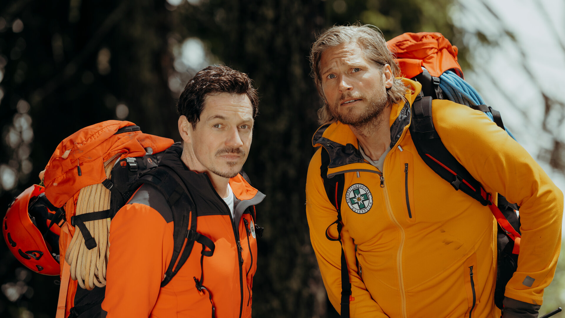 "Die Bergretter - Aus Angst":  Vor einem begrünten Berghang: Die Bergretter Markus (Sebastian Ströbel) und Tobi (Markus Brandl) stehen nebeneinander und blicken aufmerksam in die Ferne. Sie tragen Rücksäcke, an denen Seile und Helme befestigt sind.