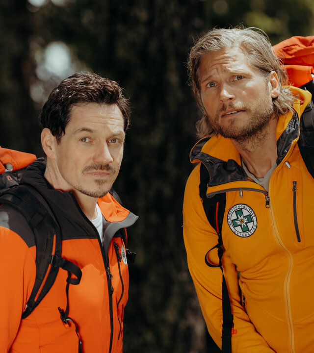 "Die Bergretter - Aus Angst":  Vor einem begrünten Berghang: Die Bergretter Markus (Sebastian Ströbel) und Tobi (Markus Brandl) stehen nebeneinander und blicken aufmerksam in die Ferne. Sie tragen Rücksäcke, an denen Seile und Helme befestigt sind.