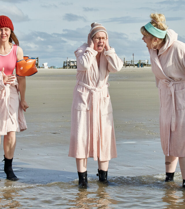 "Fast perfekte Frauen": Suse (Silke Bodenbender), Christine (Anne Roemeth), Doris (Tanja Schleiff), Jennifer (Malene Becker) und Leisha (Sabrina Amali) stehen mit hellem Bademantel, Mützen und Stiefeln im Meer. Um sie herum ist nur weiter Sandstrand und sonst keine Menschenseele. Während Suse, Doris und Jennifer laut vor sich hin schreien, zeigen Christine und Leisha eher verhaltene Reaktionen.