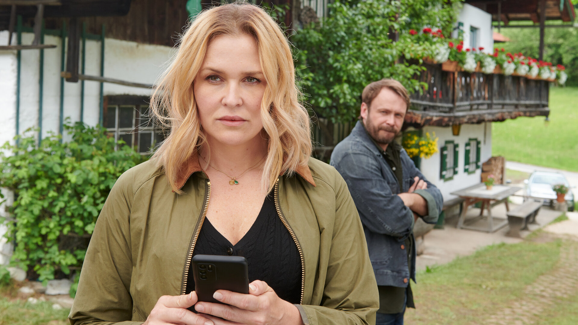 "Lena Lorenz - Schicksalsschlag": Vor dem Lorenzhof stehen Lena Lorenz (Judith Hoersch) und Dr. Aaron Kolberg (Matthias Ziesing). Lena steht im Vordergrund, hält ein Handy in den Händen und blickt nachdenklich. Im Hintergrund wird sie dabei von Aaron beobachtet.