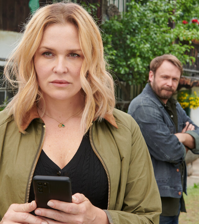 "Lena Lorenz - Schicksalsschlag": Vor dem Lorenzhof stehen Lena Lorenz (Judith Hoersch) und Dr. Aaron Kolberg (Matthias Ziesing). Lena steht im Vordergrund, hält ein Handy in den Händen und blickt nachdenklich. Im Hintergrund wird sie dabei von Aaron beobachtet.