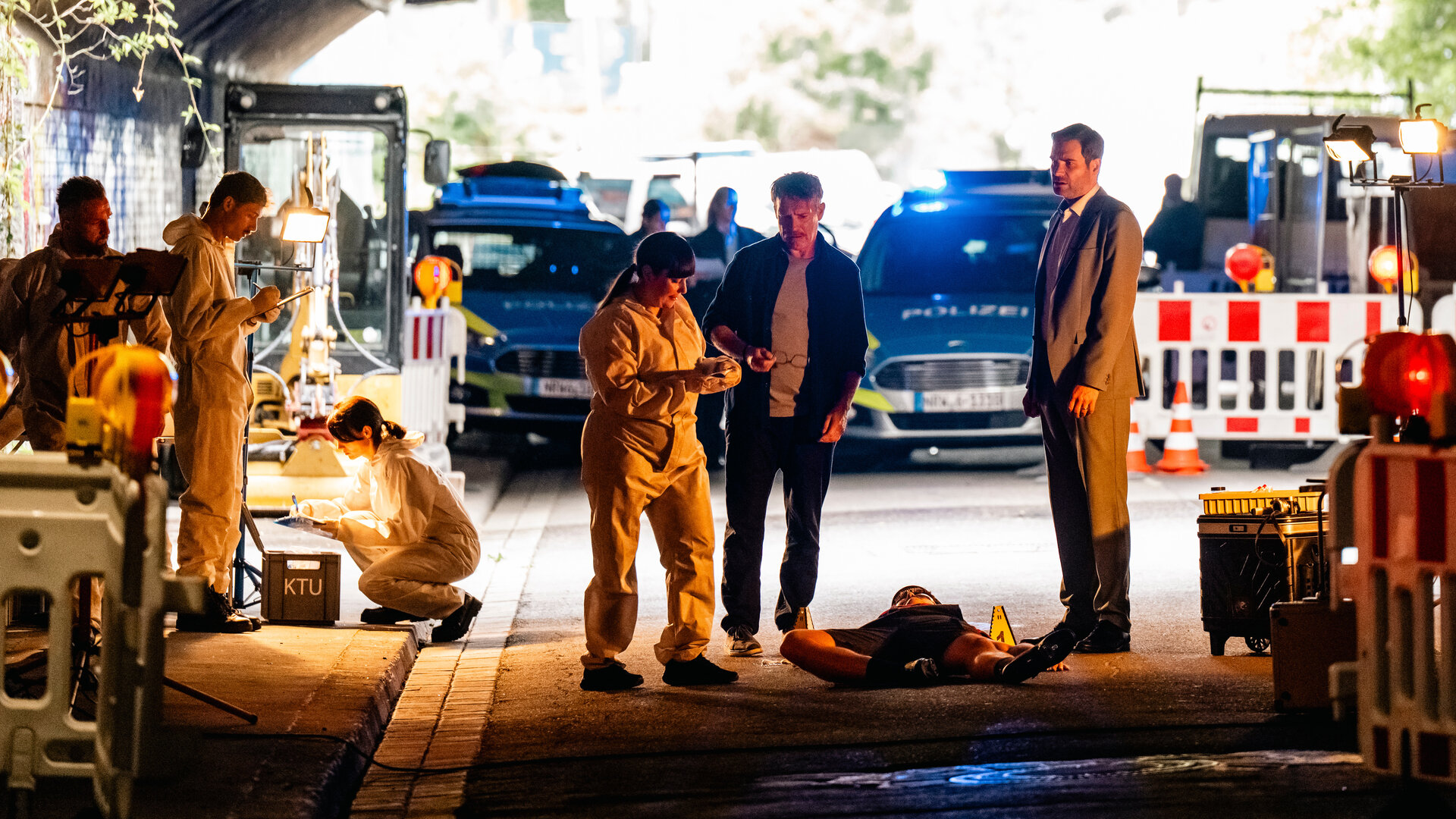 "SOKO Köln - Straßenkatzen": Matti Wagner (Pierre Besson) und Jonas Fischer (Lukas Piloty) stehen neben Dr. Pilar Westphal (Yulia Yáñez Schmidt) in einer Unterführung. Vor ihnen liegt auf dem Rücken der Tote Ole Segg (Daniel Peukmann). Die beiden Kommissare blicken zur Rechtsmedizinerin. Diese sieht auf einen Gegenstand, den sie in den Händen hält. Sie trägt einen weißen Spurensicherungsanzug. Hinter ihr arbeiten drei weitere Mitarbeiter (Komparsen) der Spurensicherung. Im Hintergrund stehen zwei Polizeiautos hinter einer rot-weiße Straßenabsperrung.
