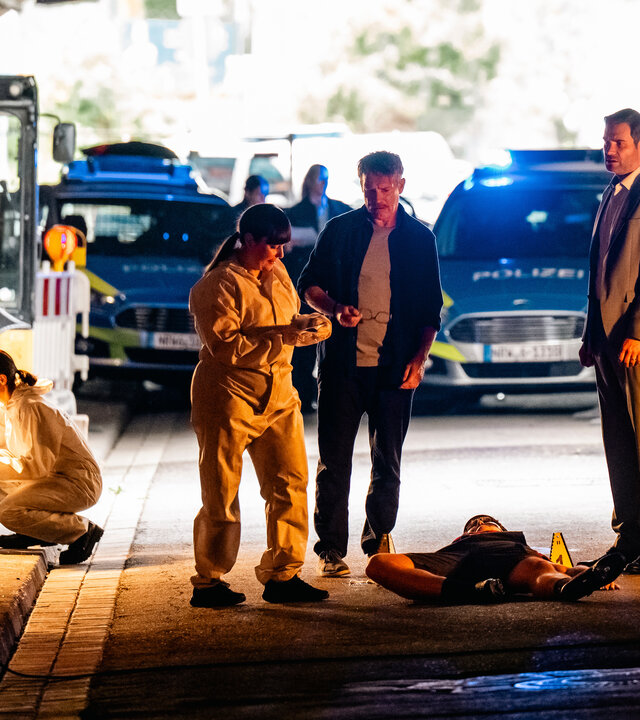 "SOKO Köln - Straßenkatzen": Matti Wagner (Pierre Besson) und Jonas Fischer (Lukas Piloty) stehen neben Dr. Pilar Westphal (Yulia Yáñez Schmidt) in einer Unterführung. Vor ihnen liegt auf dem Rücken der Tote Ole Segg (Daniel Peukmann). Die beiden Kommissare blicken zur Rechtsmedizinerin. Diese sieht auf einen Gegenstand, den sie in den Händen hält. Sie trägt einen weißen Spurensicherungsanzug. Hinter ihr arbeiten drei weitere Mitarbeiter (Komparsen) der Spurensicherung. Im Hintergrund stehen zwei Polizeiautos hinter einer rot-weiße Straßenabsperrung.
