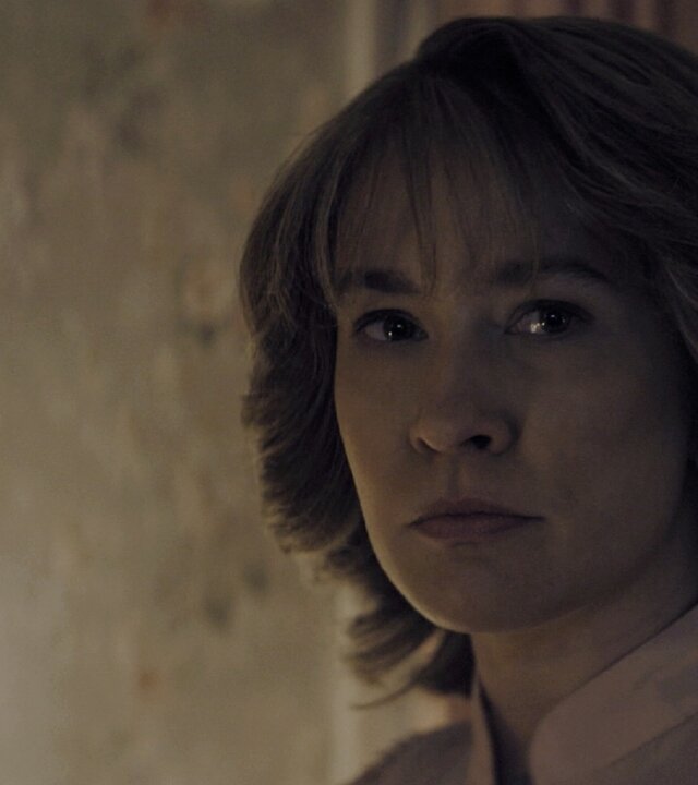 "Hysteria! - Leben aus dem Tod": Tracy (Anna Camp) steht in einem schwach beleuchteten Raum und blickt ernst zur Seite. Neben ihr hängt ein Kruzifix an der Wand.