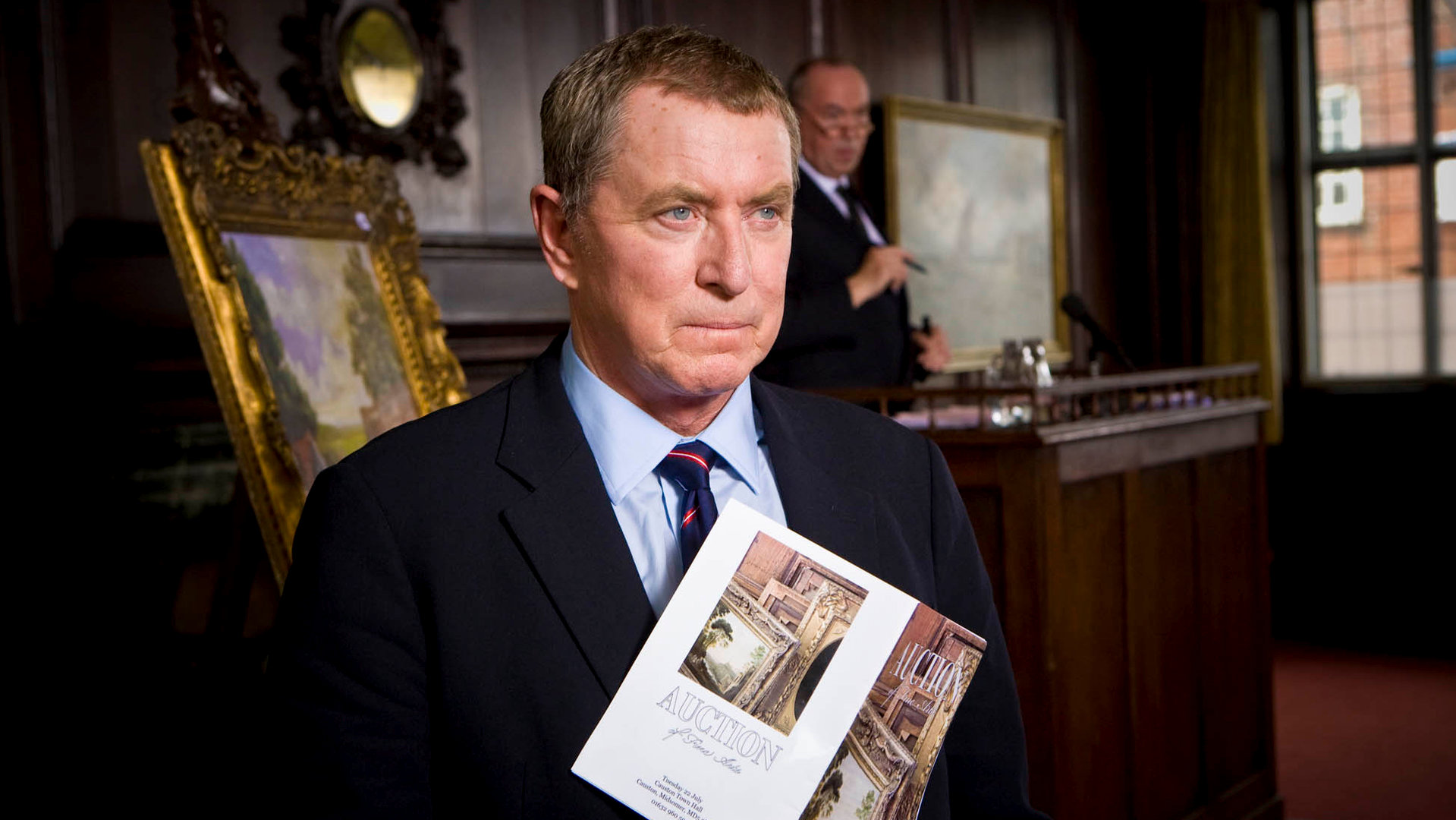 "Inspector Barnaby - Morden ist auch eine Kunst": Nahaufnahme von DCI Tom Barnaby (John Nettles) mit einem Auktionskatalog in der Hand. Hinter ihm steht ein Auktionator an seinem Pult.