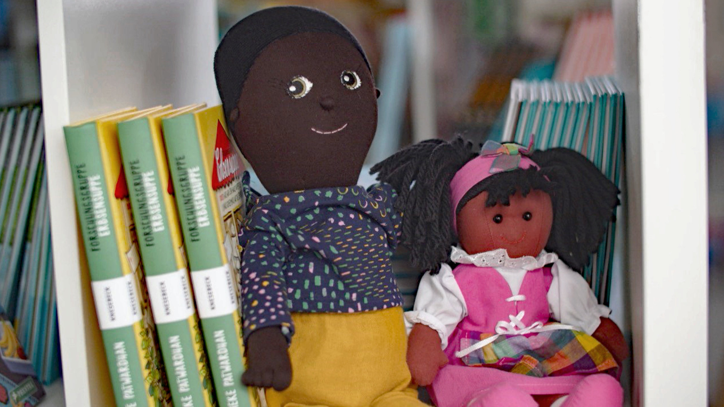 "Woher kommst du eigentlich? Schwarze in Deutschland": Zwei Kinderpuppen sitzen in einem Regal zwischen Büchern.