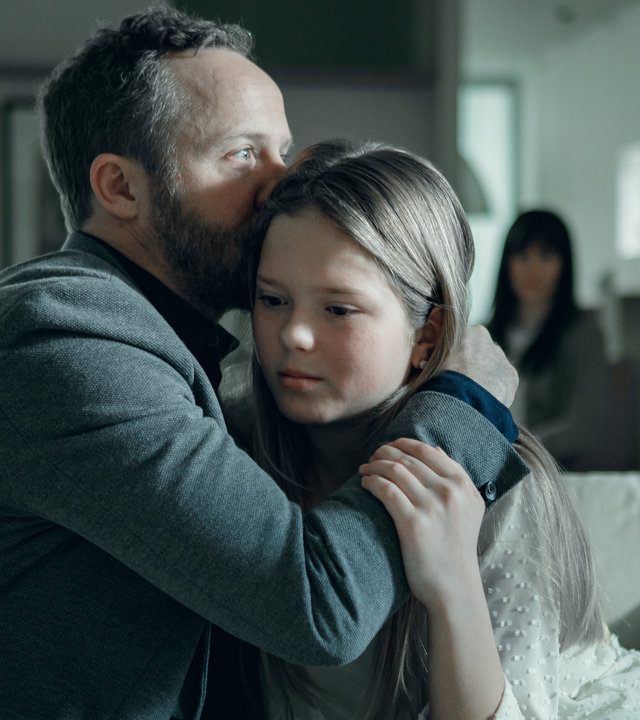 "Arctic Circle - Der unsichtbare Tod (7)": Dr. Thomas Lorenz (Maximilian Brückner) sitzt mit seiner Tochter Hanna (Lilli Salonen) auf einem Sofa. Er küsst ihr Haar. Im Hintergrund sitzt ihre Mutter Gunilla (Maria Ylipää).