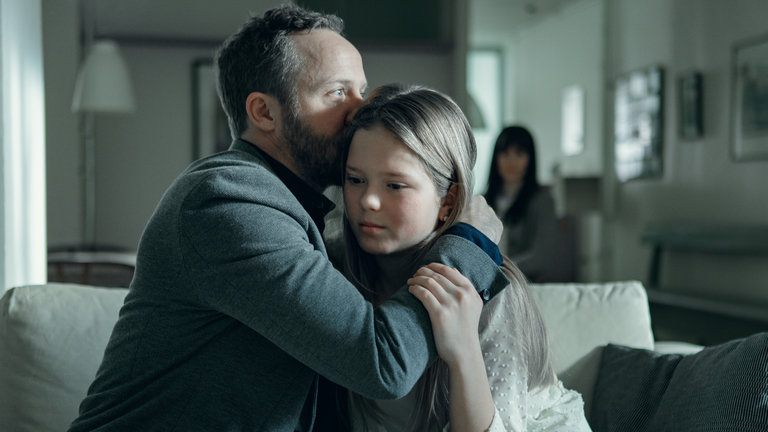 "Arctic Circle - Der unsichtbare Tod (7)": Dr. Thomas Lorenz (Maximilian Brückner) sitzt mit seiner Tochter Hanna (Lilli Salonen) auf einem Sofa. Er küsst ihr Haar. Im Hintergrund sitzt ihre Mutter Gunilla (Maria Ylipää).