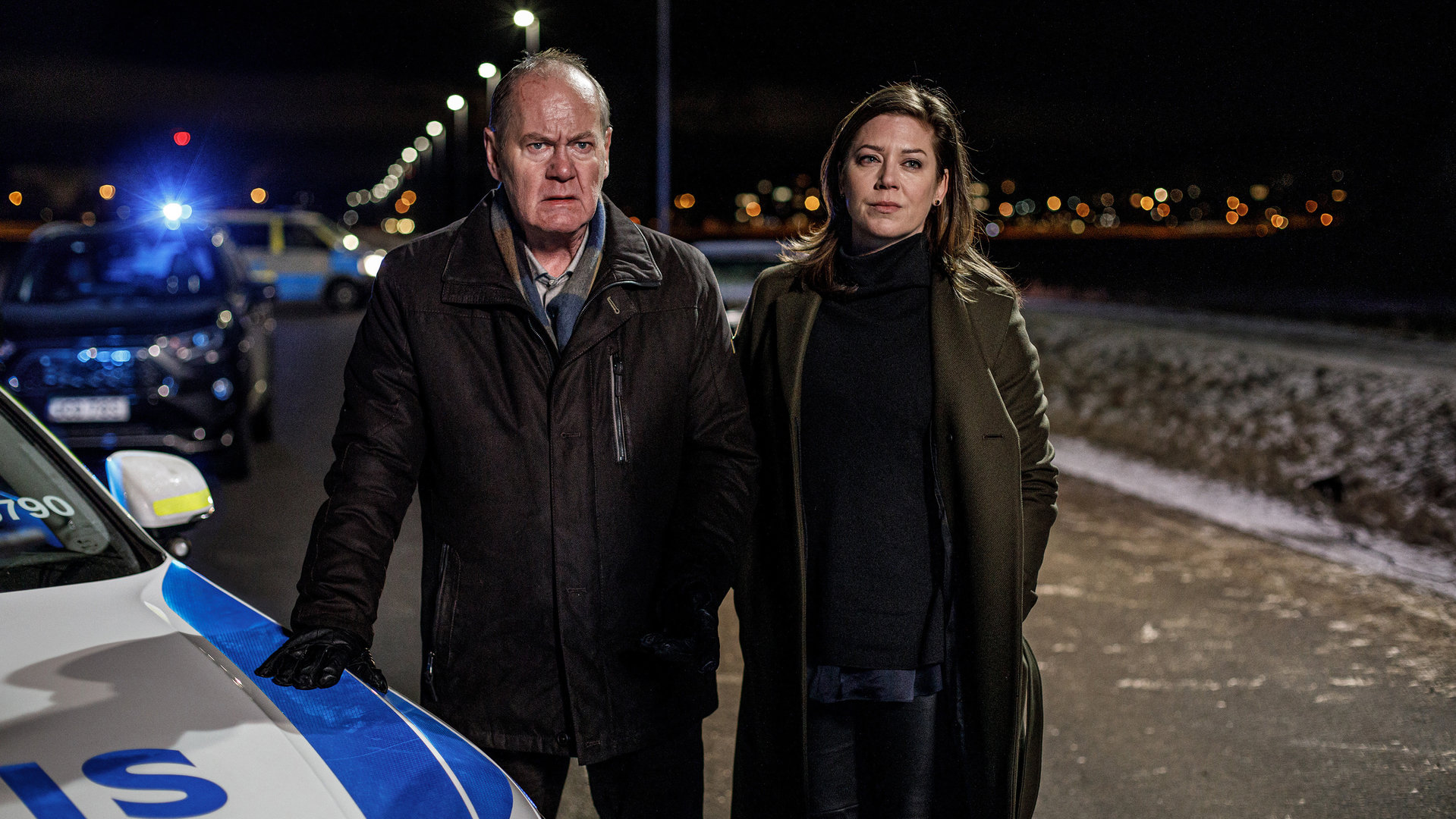 "Kommissar Beck: Die betroffene Polizei": In der Dunkelheit stehen Martin Beck (Peter Haber) und Alex Beijer (Jennie Silfverhjelm) besorgt auf einer Landstraße neben einem Polizeiauto. Hinter ihnen sind weitere Einsatzfahrzeuge der Polizei zu sehen.