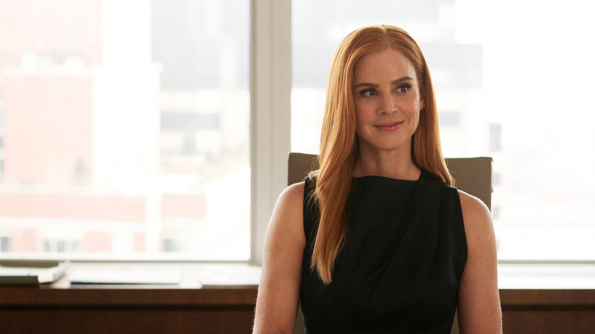 "Suits - Wem der Schuh passt": Donna (Sarah Rafferty) sitzt in einem eleganten schwarzen Top an einem Schreibtisch, ihre Hände ruhen dabei auf dem Tisch. Sie schaut glücklich und lächelt dabei und trägt ihre roten Haare offen.