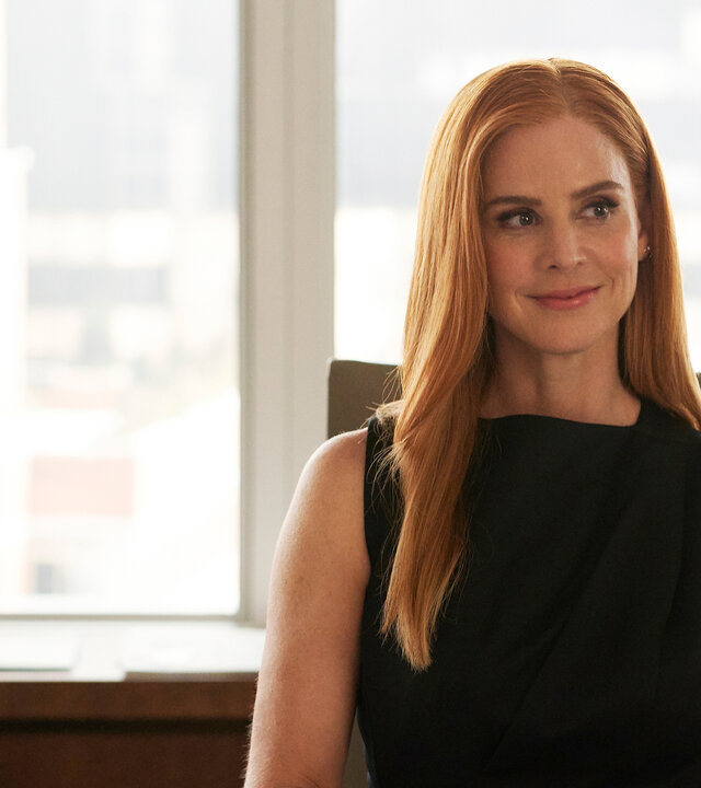 "Suits - Wem der Schuh passt": Donna (Sarah Rafferty) sitzt in einem eleganten schwarzen Top an einem Schreibtisch, ihre Hände ruhen dabei auf dem Tisch. Sie schaut glücklich und lächelt dabei und trägt ihre roten Haare offen.