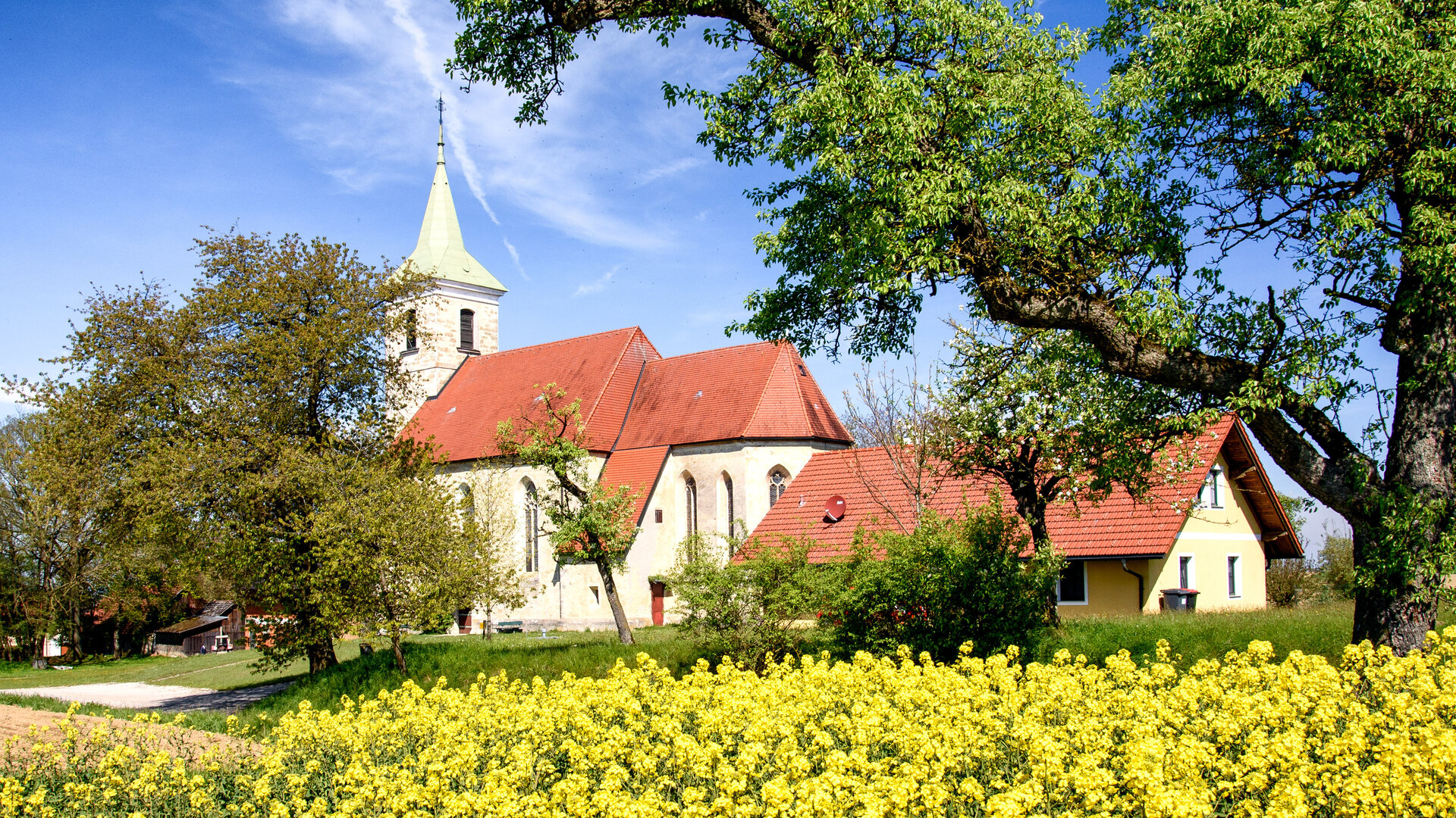 "Katholischer Gottesdienst - Unterwegs als Hörende": Die Filialkirche im oberösterreichischen Weigersdorf liegt idyllisch im Grünen. Im Vordergrund blüht leuchtend gelb der Raps, dahinter befindet sich die kleine Kirche aus hellem Stein mit rotem Dach, umgeben von Bäumen. Der eckige Kirchturm ragt in den blauen Himmel.