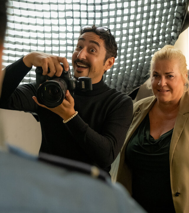 "Blutige Anfänger - Vermisste Schwester": Rico (Eko Fresh) und Dörte (Petra Kleinert) stehen dicht nebeneinander in einem Raum. Sie grinsen zu Phillip (Eric Cordes), der ihnen mit dem Rücken zur Kamera gegenübersteht. Rico hält eine Kamera in den Händen.
