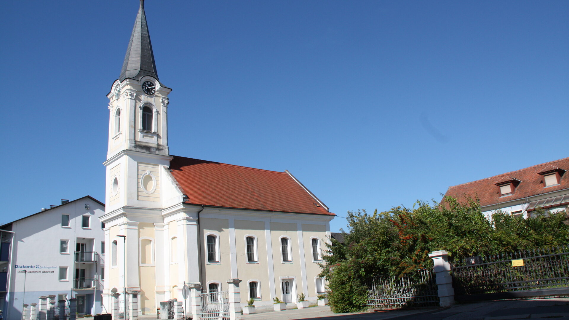 "Evangelischer Gottesdienst - Schau, es geht doch!": Die Pfarrkirche Oberwart im österreichischen Südburgenland ist außen hellgelb-weiß gestrichen, ihr Dach hat rote Ziegel. Der mit Schiefer bedeckte, eckige Kirchturm ragt in den wolkenlosen, blauen Himmel.