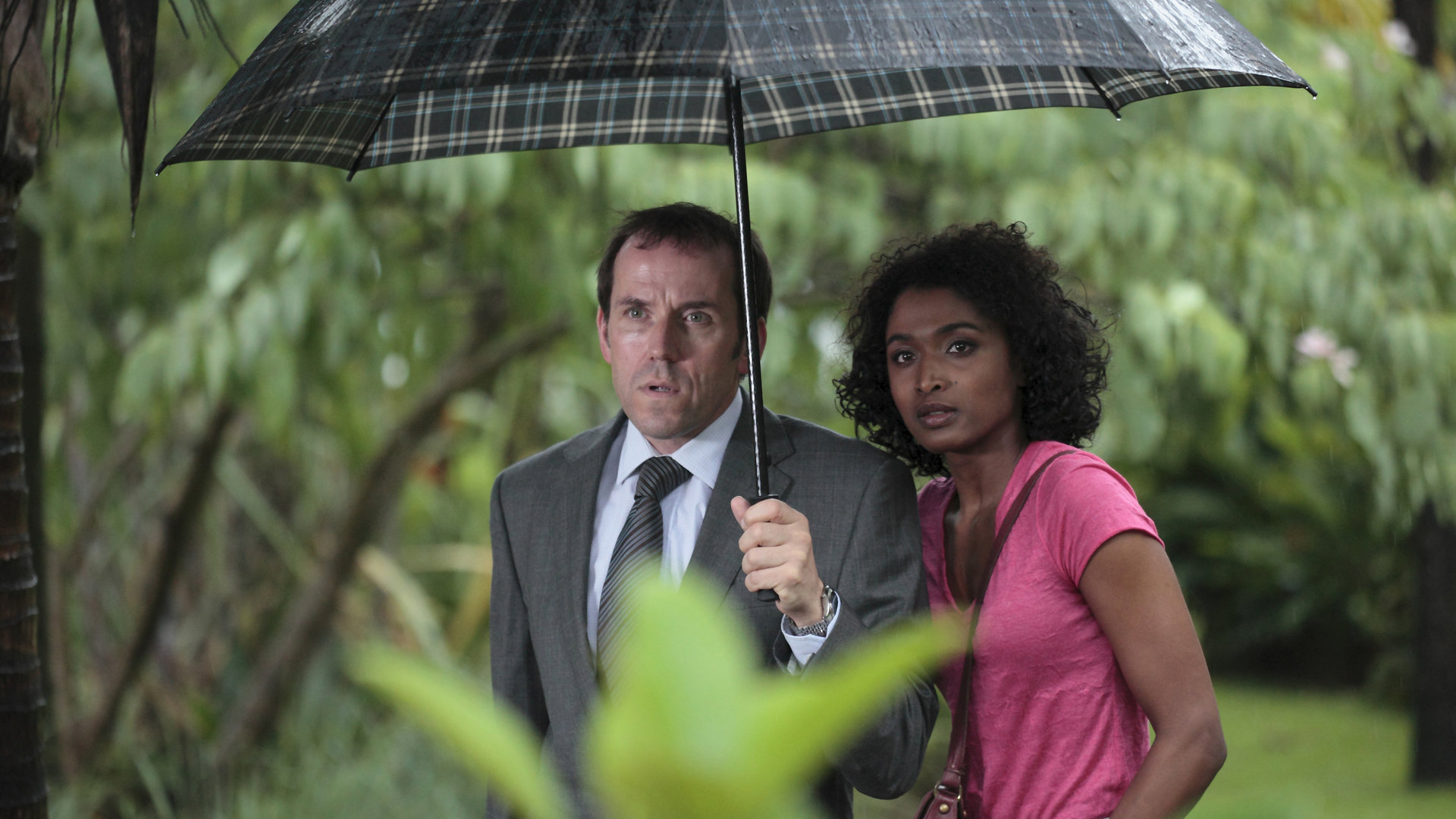 "Death in Paradise - Falsche Töne": Außenaufnahme DI Richard Poole (Ben Miller) und Camille Bordey (Sara Martins) - beide stehen unter einem Regenschirm, der von Poole gehalten wird.