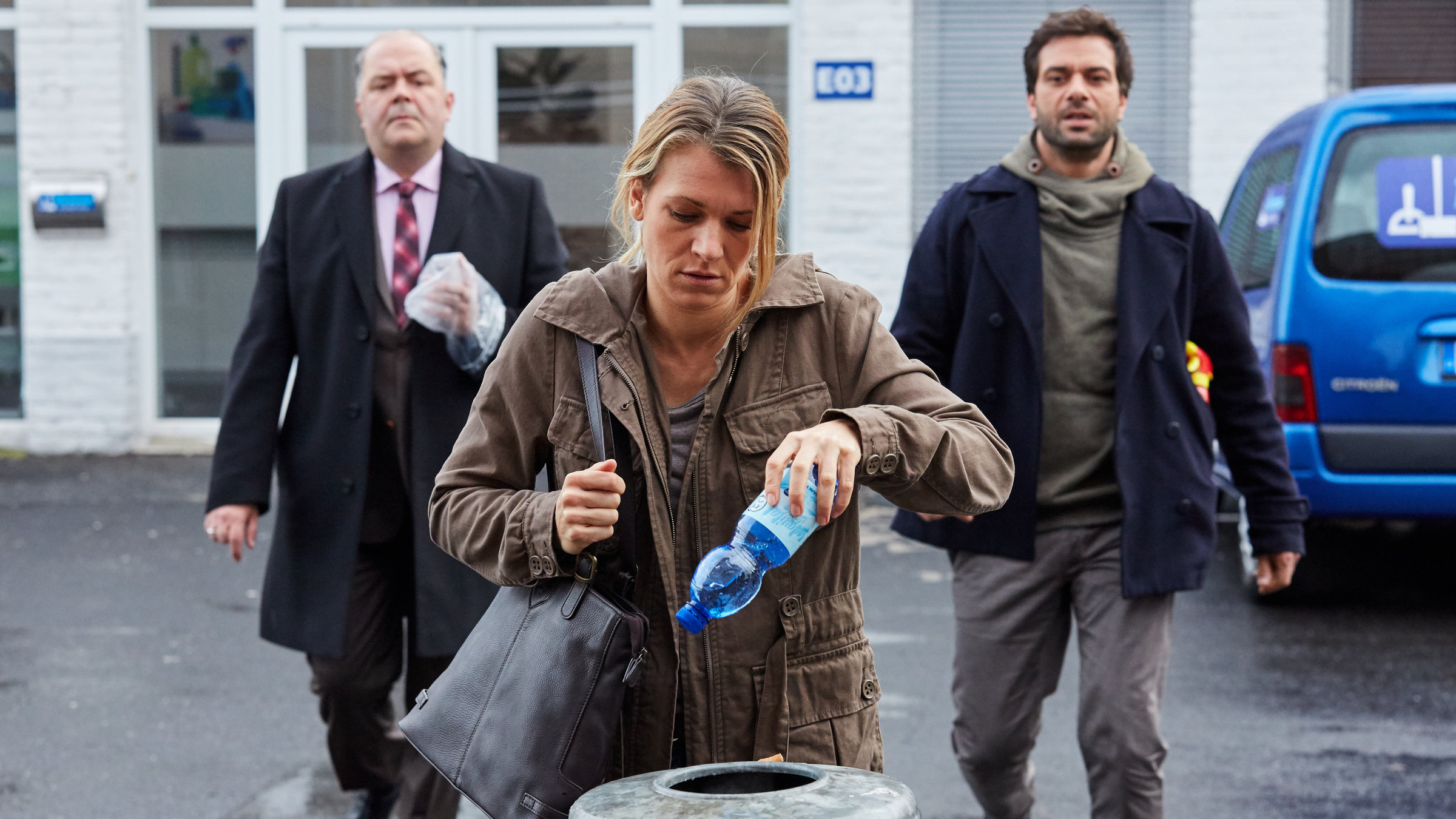 "Heldt - Der Putzteufel": Anja Reberlein (Kerstin Landsmann) steht vor dem Abfalleimer eines Hauses und will eine PET-Flasche hineinwerfen. Hinter ihr kommen Grün (Timo Dierkes) und Heldt (Kai Schumann) angelaufen.