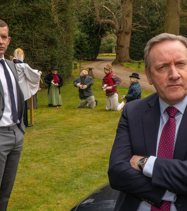 "Inspector Barnaby - Die Vogelscheuchen-Morde": DS Jamie Winter (Nick Hendrix) steht auf einer Wiese, auf der fünf Vogelscheuchen zu sehen sind. Im vorderen Teil des Bildes lehnt DCI John Barnaby (Neil Dudgeon) mit verschränkten Armen auf der Motorhaube eines Wagens und sieht in die Kamera.