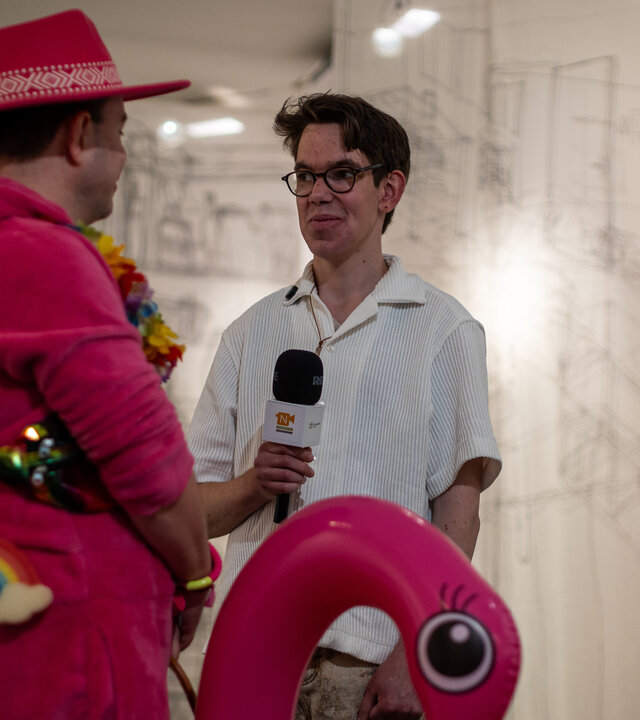 "einfach Mensch - Justus: Ich bin Reporter": Justus steht in einer Galerie mit einem Mikrofon in der Hand neben der queeren Kunstfigur Flirty Flamingo.