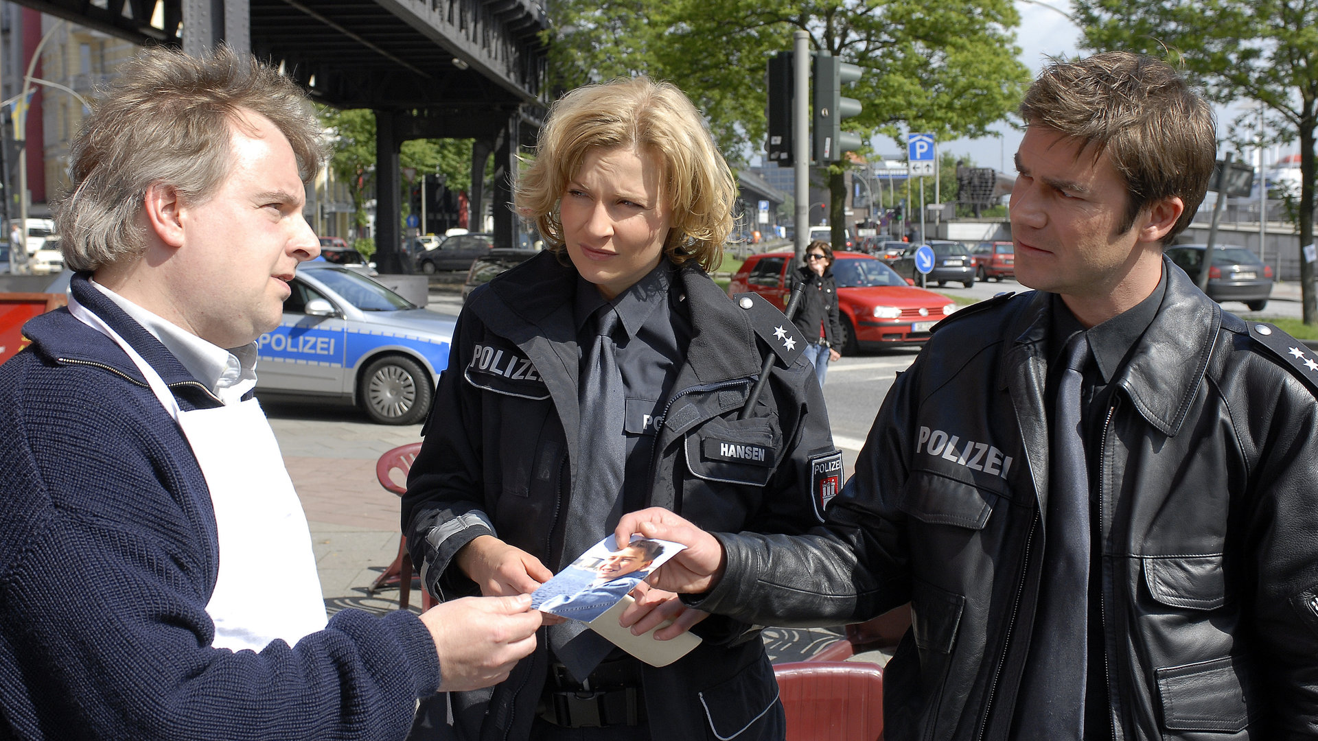 "Notruf Hafenkante - Das schwarze Kleid": Nils (Thomas Scharff) und Melanie (Sanna Englund) stehen zusammen mit dem Imbissbudenbesitzer (Rüdiger Dambroth) auf einer Straße und befragen ihn. Nils zeigt ihm ein Foto.