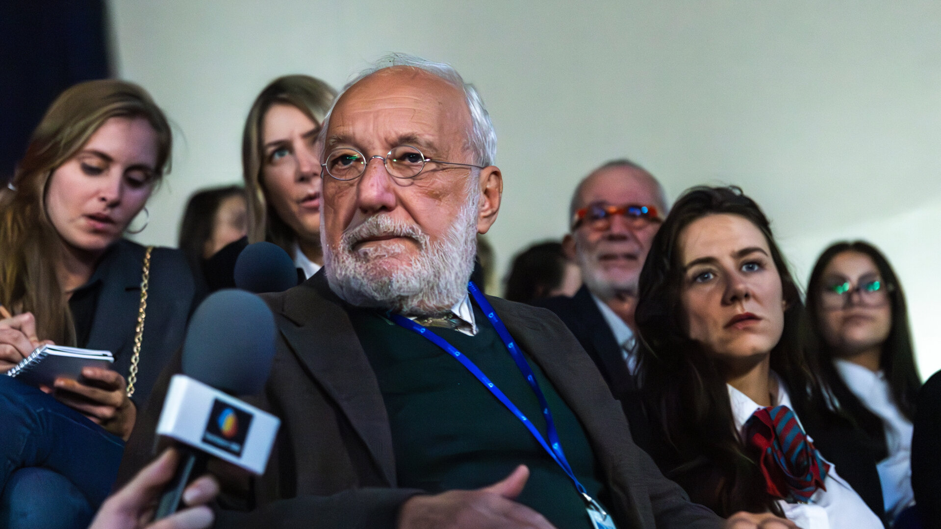 "Phoenix - Change Or Die: 12 Milliarden Euro": Professor Jean Humbel (François Berléand, M.) hat einen grauen Vollbart und schütteres weißes Haar. Er sitzt auf einem Klappsitz eines vollbesetzten Auditoriums und hat seinen Kopf zu einem Reporter (Komparse) gedreht, der neben ihm sitzt und ihm ein Mikrofon entgegenhält.