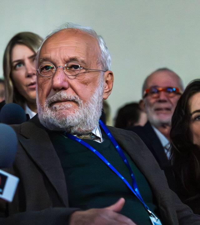 "Phoenix - Change Or Die: 12 Milliarden Euro": Professor Jean Humbel (François Berléand, M.) hat einen grauen Vollbart und schütteres weißes Haar. Er sitzt auf einem Klappsitz eines vollbesetzten Auditoriums und hat seinen Kopf zu einem Reporter (Komparse) gedreht, der neben ihm sitzt und ihm ein Mikrofon entgegenhält.