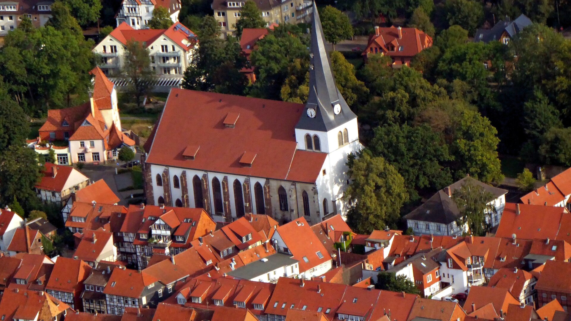 Evangelischer Gottesdienst - "Lehn dich einfach an": Blick von schräg oben auf die Kirche St. Sixti in Northeim: Die weiße Kirche mit rotem Dach und einem spitz zulaufenden Kirchturm liegt inmitten der Altstadt und ist umgeben von Fachwerkhäusern und von Bäumen.