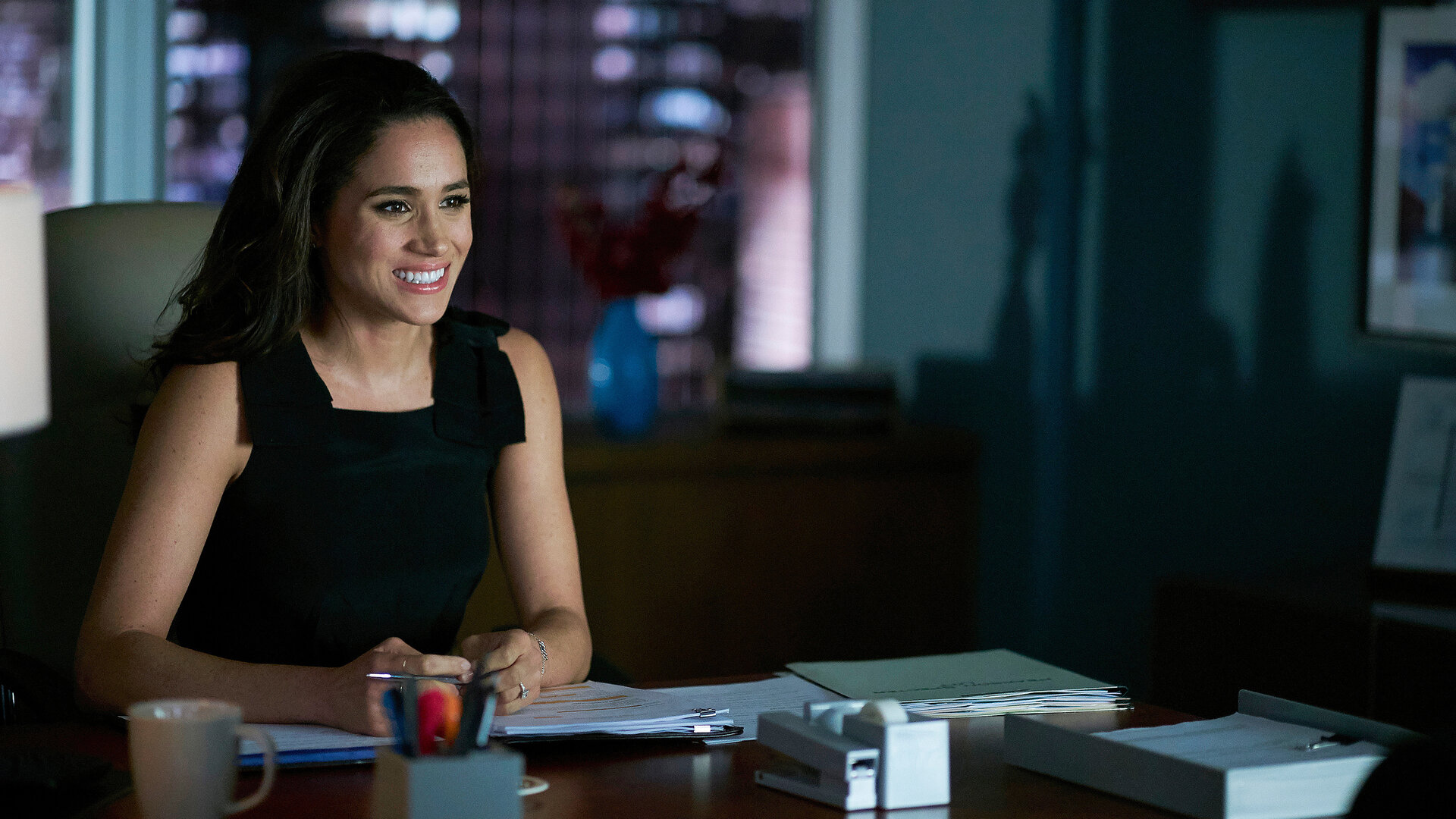 "Suits - Zwietracht": Rachel sitzt am Abend im dunklen Büro am Schreibtisch. Sie hält einen Stift in den Händen, die auf Dokumenten liegen. Sie lächelt und sieht erleichtert aus.