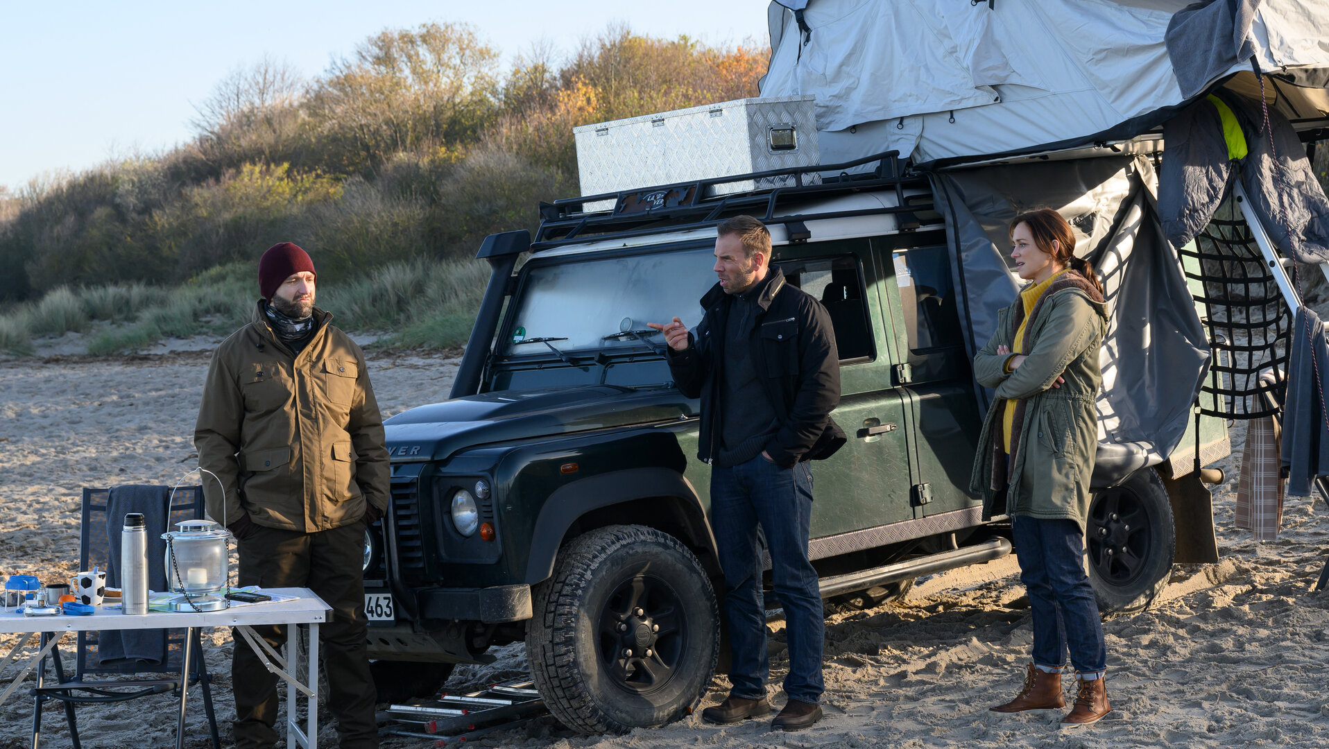 "SOKO Wismar - Schatzinsel Poel": Am Strand steht ein Auto mit Zeltaufbau, daneben ein Campingtisch. Davor wird Darius Kopp (Aaron Thiesse) von Karoline Joost (Nike Fuhrmann) und Lars Pöhlmann (Dominic Boeer) befragt.