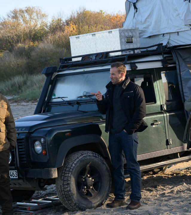 "SOKO Wismar - Schatzinsel Poel": Am Strand steht ein Auto mit Zeltaufbau, daneben ein Campingtisch. Davor wird Darius Kopp (Aaron Thiesse) von Karoline Joost (Nike Fuhrmann) und Lars Pöhlmann (Dominic Boeer) befragt.