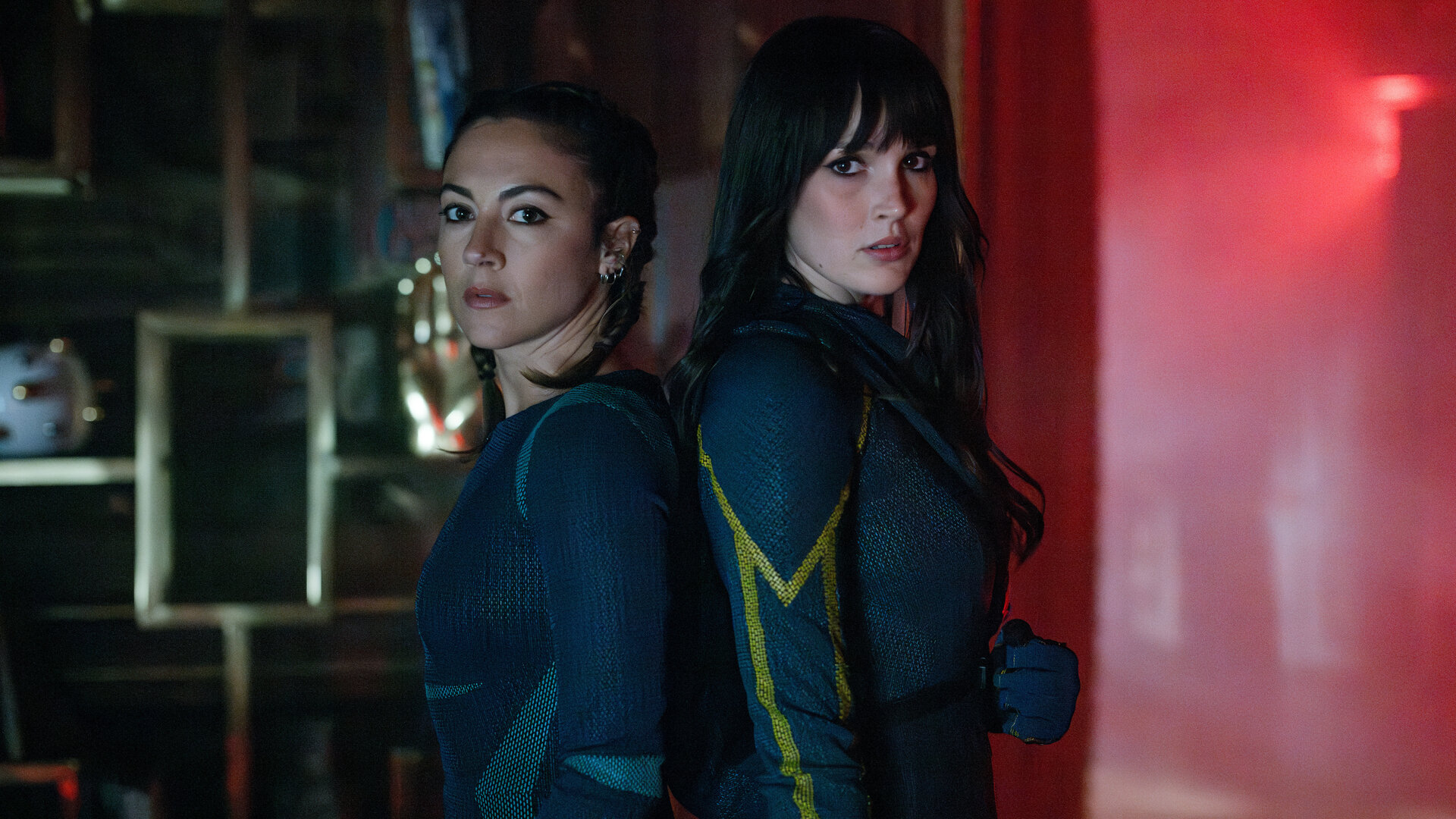 "Cat's Eyes - Heinz": Sylia (Constance Labbé) und Tamara (Camille Lou) stehen Rücken an Rücken in einem dunklen Raum und blicken in die Kamera. Im Hintergrund leuchtet ein rotes Licht.