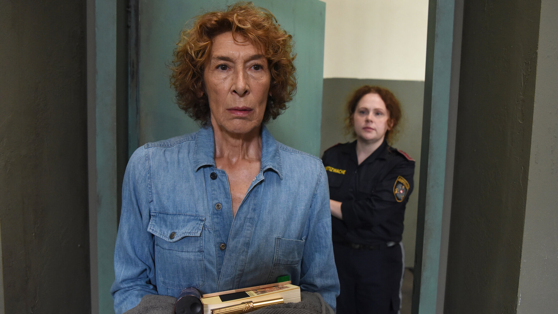 "Mama ist die Best(i)e Teil I": Gloria Almeda (Adele Neuhauser) trägt Gefängniskleidung. Sie wird von einer Justizbeamtin zu ihrer Zelle gebracht und hält einige Utensilien in Händen. Die Tür hinter ihr wird sich gleich schließen.
