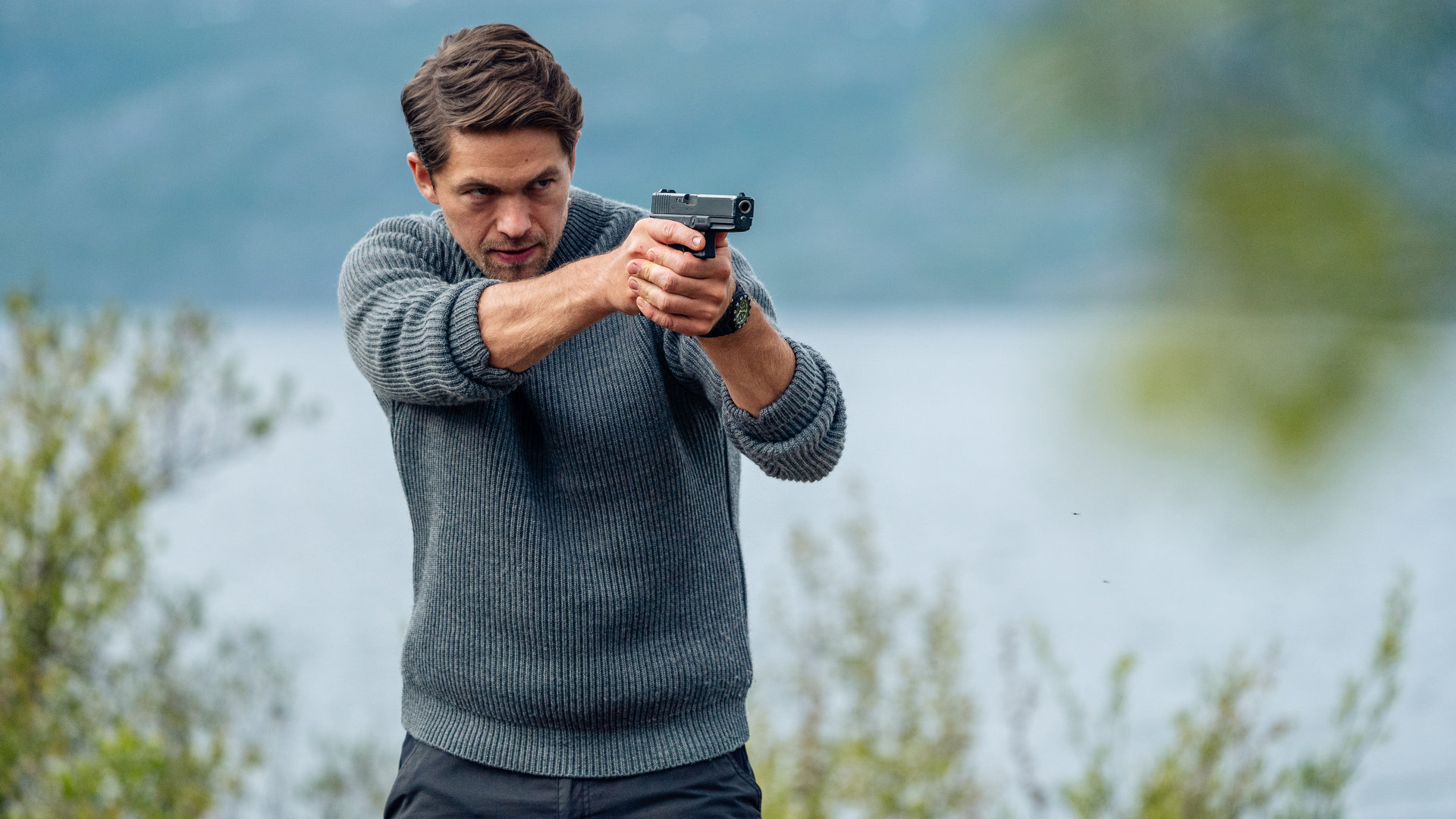 "Hamilton - Das Kiruna Projekt": Hamilton (Jakob Ofterbro) hält eine Waffe im Anschlag. Er trägt einen Wollpullover. Im Hintergrund ist ein See zu sehen.