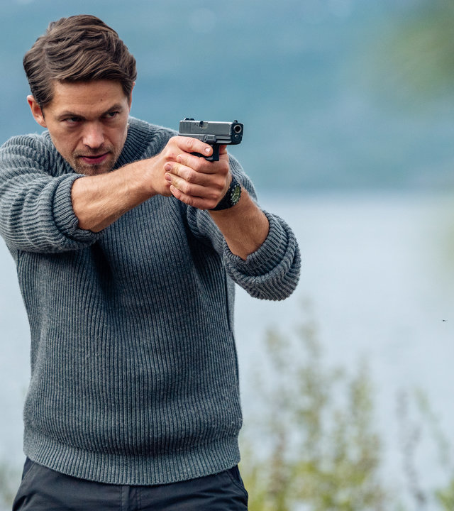 "Hamilton - Das Kiruna Projekt": Hamilton (Jakob Ofterbro) hält eine Waffe im Anschlag. Er trägt einen Wollpullover. Im Hintergrund ist ein See zu sehen.