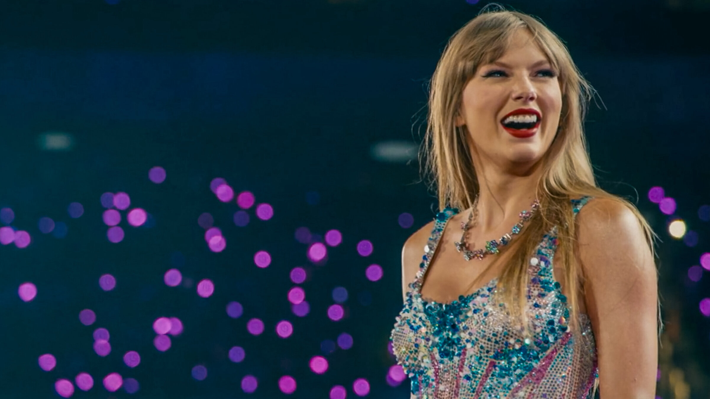 "Becoming Taylor Swift - Ruhm und Rivalen": Taylor Swift steht auf der Bühne und trägt ein glitzerndes, buntes Kleid und eine auffällige Halskette vor unscharfem Hintergrund mit lila Lichtpunkten.