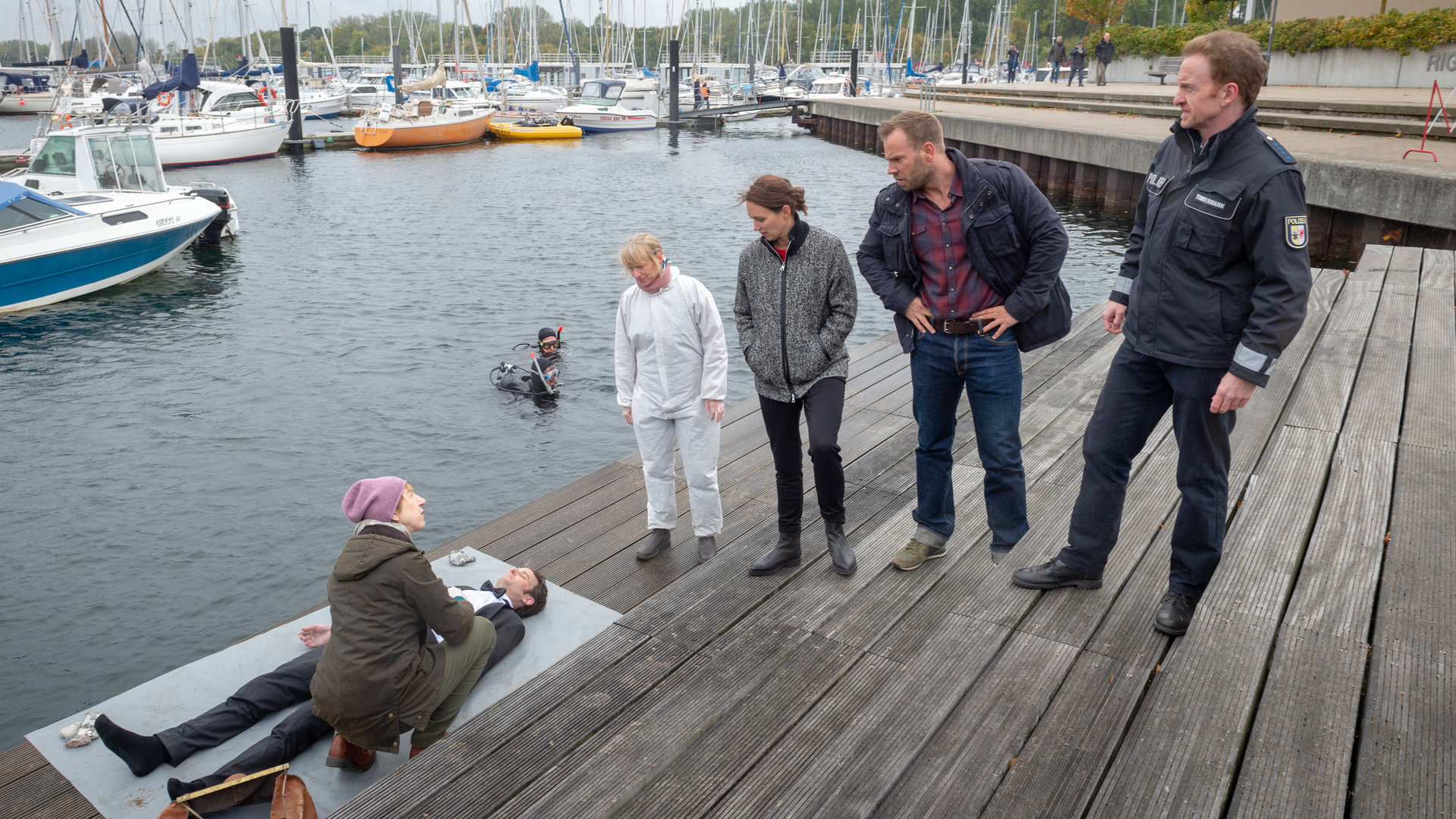 "SOKO Wismar - Mundraub":  Auf einem großzügigen Holzsteg am Yachthafen steht das Team der SOKO (Roswitha Prinzler (Silke Matthias), Karoline Joost (Nike Fuhrmann), Lars Pöhlmann (Dominic Boeer), Timmermann (Mathias Junge)) nebeneinander aufgereiht. Alle blicken zur Leiche von Dr. Johannes Urbach (Helge Gutbrod), der aus dem Wasser gefischt worden ist.  Helene Sturbeck (Katharina Blaschke) kniet bei der Leiche und blickt zu ihren Kollegen. Im Wasser liegen Segelboote und ein Taucher ist soeben aufgetaucht.