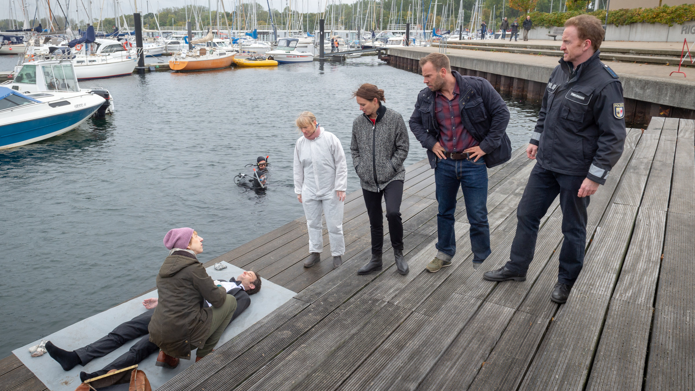 "SOKO Wismar - Mundraub":  Auf einem großzügigen Holzsteg am Yachthafen steht das Team der SOKO (Roswitha Prinzler (Silke Matthias), Karoline Joost (Nike Fuhrmann), Lars Pöhlmann (Dominic Boeer), Timmermann (Mathias Junge)) nebeneinander aufgereiht. Alle blicken zur Leiche von Dr. Johannes Urbach (Helge Gutbrod), der aus dem Wasser gefischt worden ist.  Helene Sturbeck (Katharina Blaschke) kniet bei der Leiche und blickt zu ihren Kollegen. Im Wasser liegen Segelboote und ein Taucher ist soeben aufgetaucht.