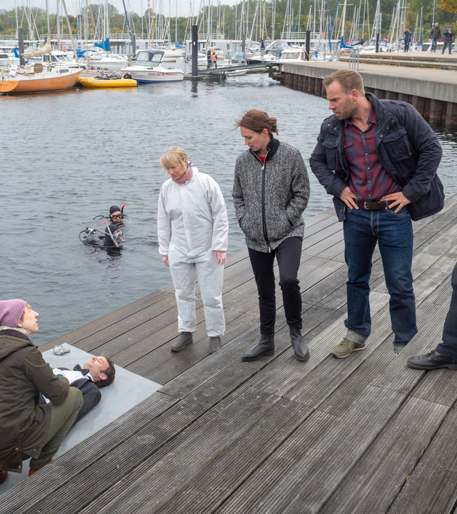 "SOKO Wismar - Mundraub":  Auf einem großzügigen Holzsteg am Yachthafen steht das Team der SOKO (Roswitha Prinzler (Silke Matthias), Karoline Joost (Nike Fuhrmann), Lars Pöhlmann (Dominic Boeer), Timmermann (Mathias Junge)) nebeneinander aufgereiht. Alle blicken zur Leiche von Dr. Johannes Urbach (Helge Gutbrod), der aus dem Wasser gefischt worden ist.  Helene Sturbeck (Katharina Blaschke) kniet bei der Leiche und blickt zu ihren Kollegen. Im Wasser liegen Segelboote und ein Taucher ist soeben aufgetaucht.