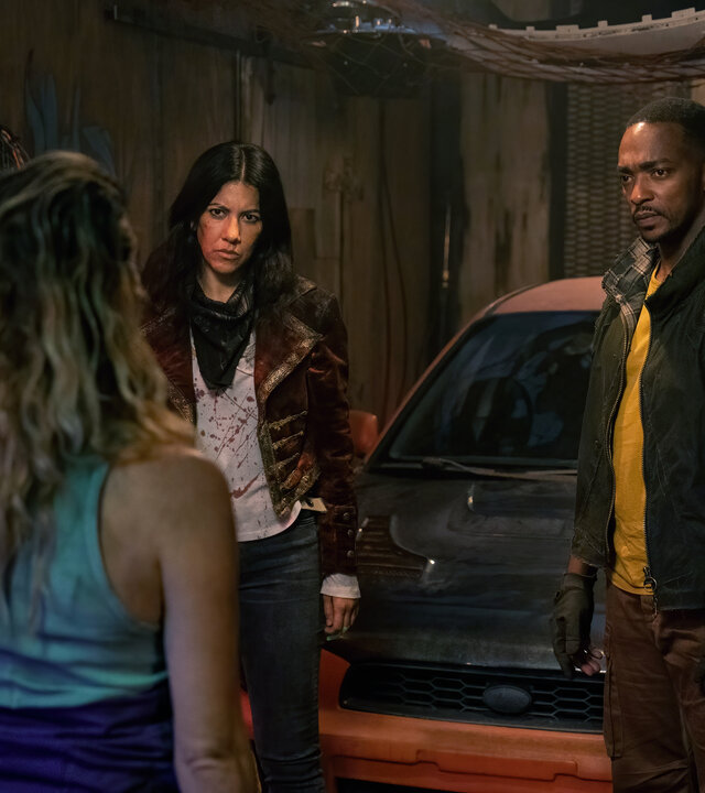 "Twisted Metal - WHZDARE": Miranda Watts (Jamie Neumann) steht in einem düsteren Raum voller Werkzeug John (Anthony Mackie) und Quiet (Stephanie Beatriz) gegenüber, die sie ernsthaft ansehen. Hinter ihnen parkt ein stark verschmutzter orangefarbener Wagen.