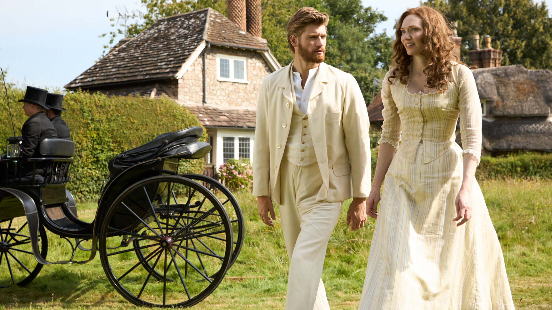 "The Forsytes - Familie verpflichtet: Krisenzeiten": Auf einer Wiese gehen Jolyon Forsyte Jr. (Danny Griffin) und Louisa (Eleanor Tomlinson) nebeneinander im Sonnenschein. Neben ihnen steht eine schwarze Kutsche mit zwei schwarz gekleideten Kutschern. Jolyon Jr. trägt einen buttergelben Anzug mit passender Weste und einem weißen Hemd darunter. Louisa trägt ein buttergelbes Kleid mit feinen Streifen und langen Ärmeln. Er blickt sie ernst an, während sie fragend nach vorn schaut. Im Hintergrund steht eine Ziegelsteinhütte mit zwei großen Schornsteinen.