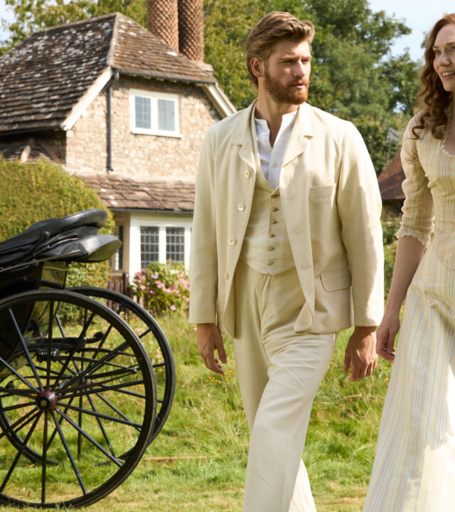 "The Forsytes - Familie verpflichtet: Krisenzeiten": Auf einer Wiese gehen Jolyon Forsyte Jr. (Danny Griffin) und Louisa (Eleanor Tomlinson) nebeneinander im Sonnenschein. Neben ihnen steht eine schwarze Kutsche mit zwei schwarz gekleideten Kutschern. Jolyon Jr. trägt einen buttergelben Anzug mit passender Weste und einem weißen Hemd darunter. Louisa trägt ein buttergelbes Kleid mit feinen Streifen und langen Ärmeln. Er blickt sie ernst an, während sie fragend nach vorn schaut. Im Hintergrund steht eine Ziegelsteinhütte mit zwei großen Schornsteinen.