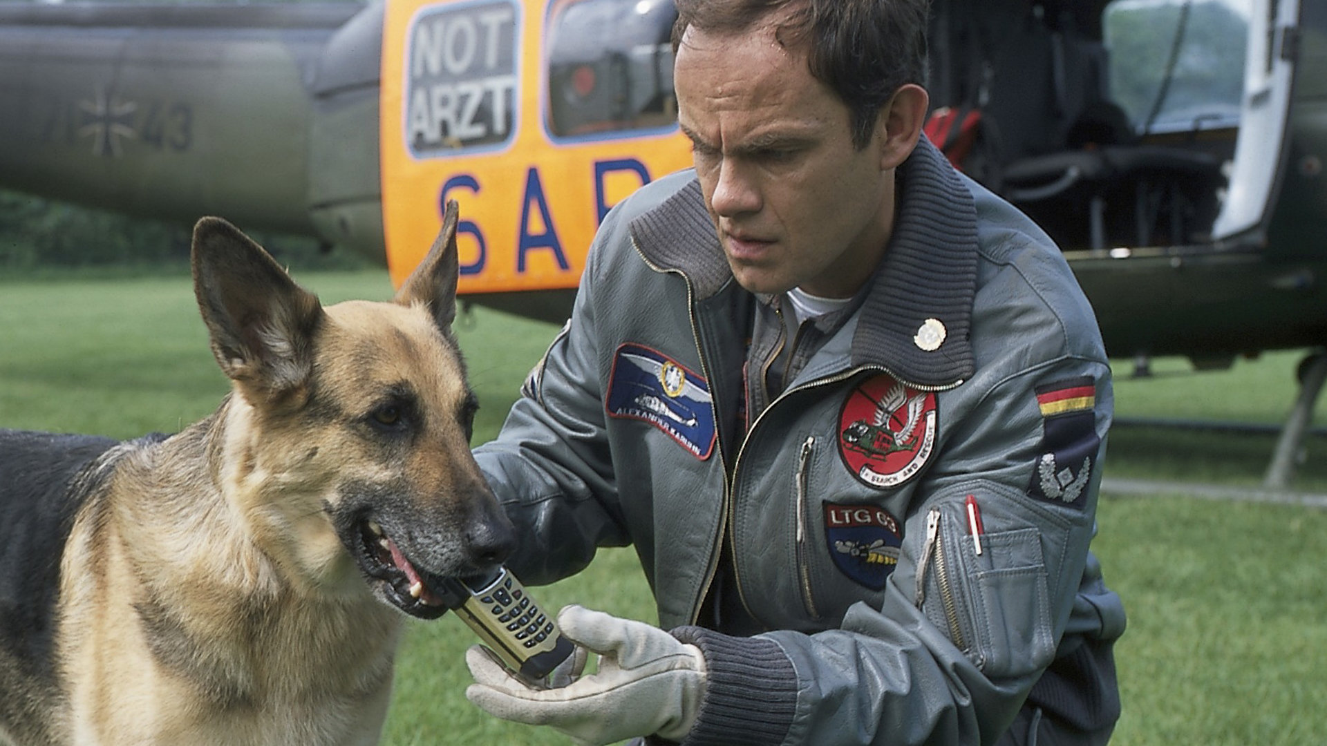 "Die Rettungsflieger - Die Zeitbombe": Alexander Karuhn (Matthias Leja) hockt auf einer Wiese neben einem Schäferhund, der ein Handy in der Schnauze trägt. Im Hintergrund ist ein Hubschrauber zu sehen.