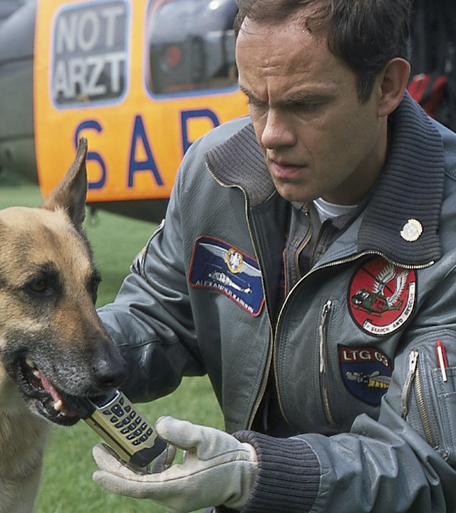 "Die Rettungsflieger - Die Zeitbombe": Alexander Karuhn (Matthias Leja) hockt auf einer Wiese neben einem Schäferhund, der ein Handy in der Schnauze trägt. Im Hintergrund ist ein Hubschrauber zu sehen.