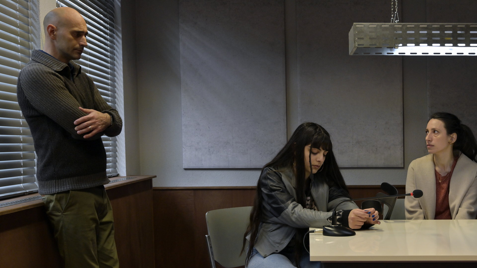 "SOKO Potsdam - Gefährliche Lügen": In einem Verhörzimmer, an einem Tisch, sitzen Jana Esen (Helena Abay) und Jessica Esen (Berfin Sönmez). Links an einem Fenster angelehnt, steht Baris Esen (Eray von Egilmez).