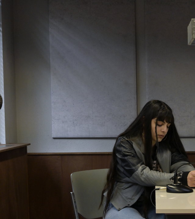 "SOKO Potsdam - Gefährliche Lügen": In einem Verhörzimmer, an einem Tisch, sitzen Jana Esen (Helena Abay) und Jessica Esen (Berfin Sönmez). Links an einem Fenster angelehnt, steht Baris Esen (Eray von Egilmez).