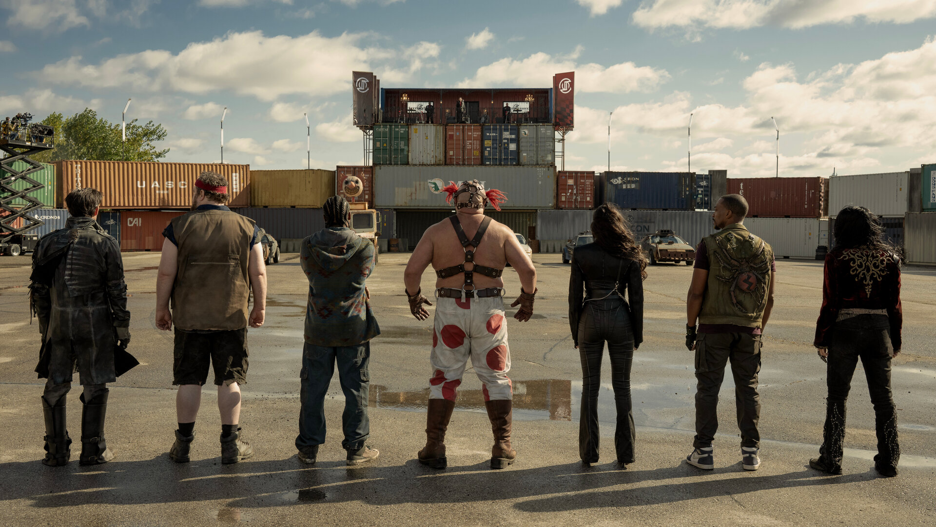 "Twisted Metal - OHLYNTE": Mr. Grimm (Richard de Klerk), Stu (Mike Mitchell), Mike (Tahj Vaughans), Sweet Tooth (Joe Seanoa), Raven (Patty Guggenheim), John (Anthony Mackie) und Quiet (Stephanie Beatriz) stehen nebeneinander auf einem Gelände vor bunten Containern. Alle stehen mit dem Rücken zur Kamera und blicken gemeinsam auf die Containerlandschaft vor ihnen.