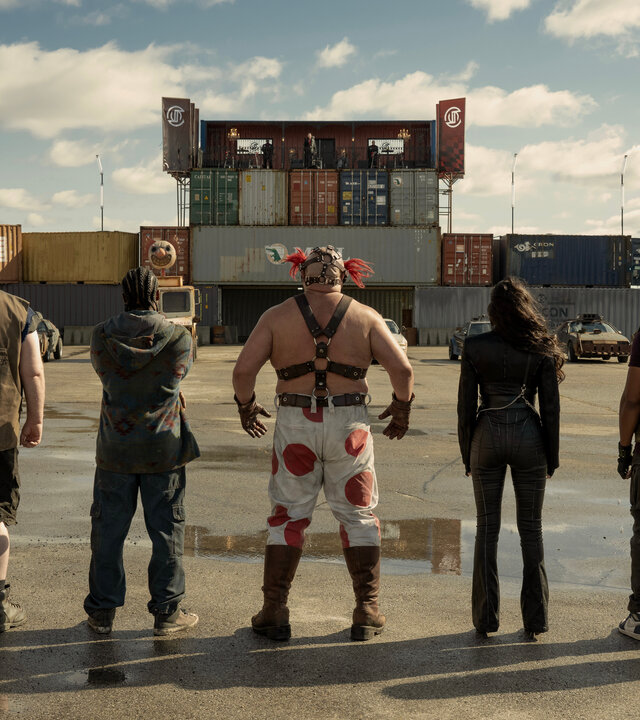 "Twisted Metal - OHLYNTE": Mr. Grimm (Richard de Klerk), Stu (Mike Mitchell), Mike (Tahj Vaughans), Sweet Tooth (Joe Seanoa), Raven (Patty Guggenheim), John (Anthony Mackie) und Quiet (Stephanie Beatriz) stehen nebeneinander auf einem Gelände vor bunten Containern. Alle stehen mit dem Rücken zur Kamera und blicken gemeinsam auf die Containerlandschaft vor ihnen.