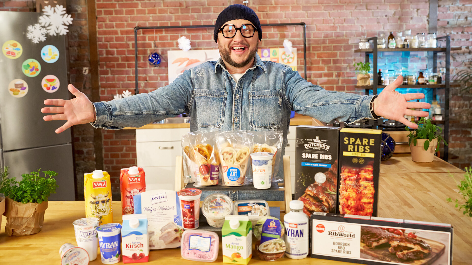 "besseresser: Klägliche Kühlprodukte - Sebastian Lege packt aus": Sebastian Lege steht hinter einem Tisch auf dem Kühlprodukte wie Ayran, Wraps, Spareribs und Matjessalat liegen.