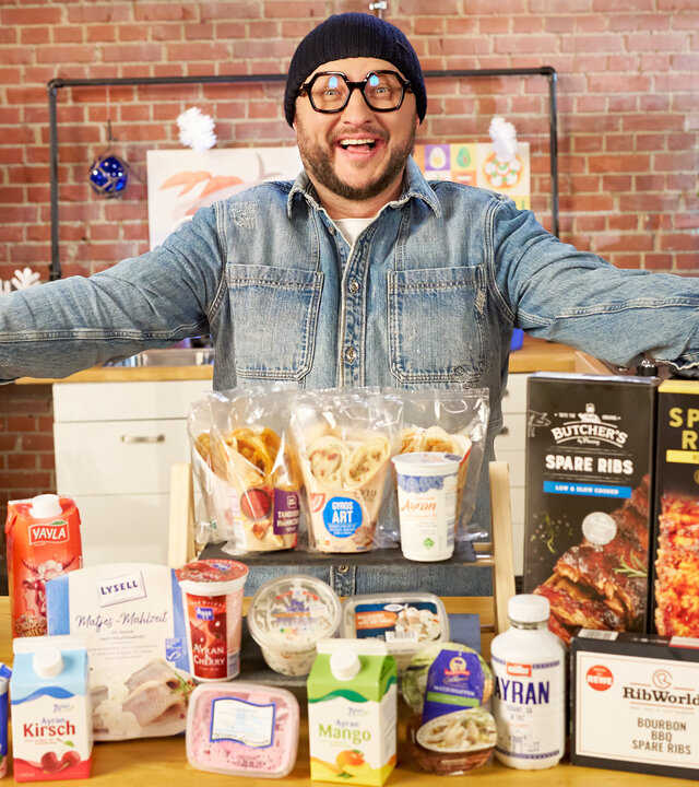 "besseresser: Klägliche Kühlprodukte - Sebastian Lege packt aus": Sebastian Lege steht hinter einem Tisch auf dem Kühlprodukte wie Ayran, Wraps, Spareribs und Matjessalat liegen.