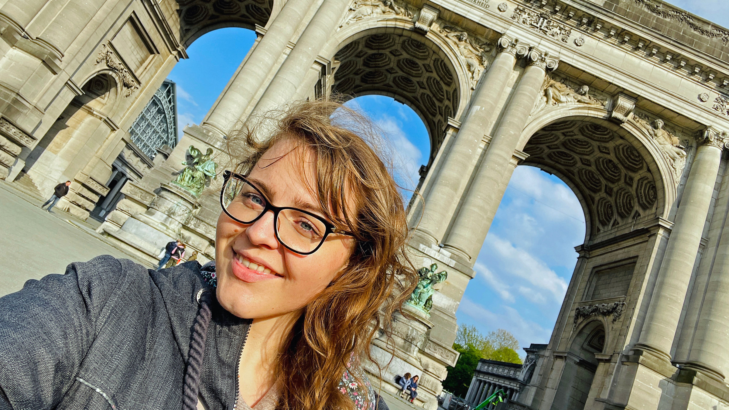 "Terra Xpress: Mikroabenteuer oder das Glück allein? Urlaub mal anders": Julia Decker mit blonden, Haaren und Brille steht lächelnd vor einem großen historischen Gebäude mit Torbögen.